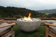 An outdoor gas fire pit named 'Infinite' with a Travertine finish, surrounded by seating areas, with a scenic landscape in the background.