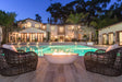 An outdoor fire bowl with a modern design, situated between two wicker chairs, with a illuminated house and pool in the background at dusk.