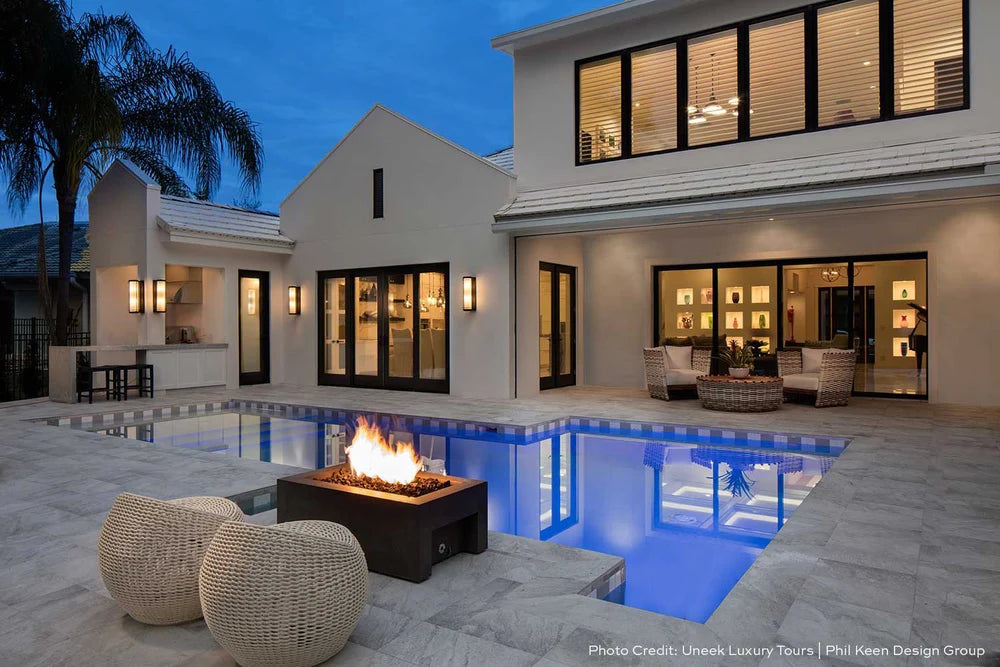 An outdoor swimming poolside area featuring a modern linear design fire pit with a gently sloped fire feature, sharp 90-degree inset walls, and surrounded by white cushions on wicker furniture.