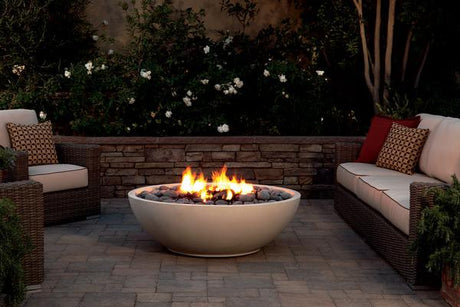 Night garden setting with lit fire pit surrounded by wicker outdoor furniture and a stone wall with concrete pavers
