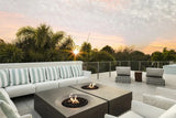 Luxury rooftop patio with two square gas fire pits, surrounded by white cushioned lounge seating and palm trees at sunset