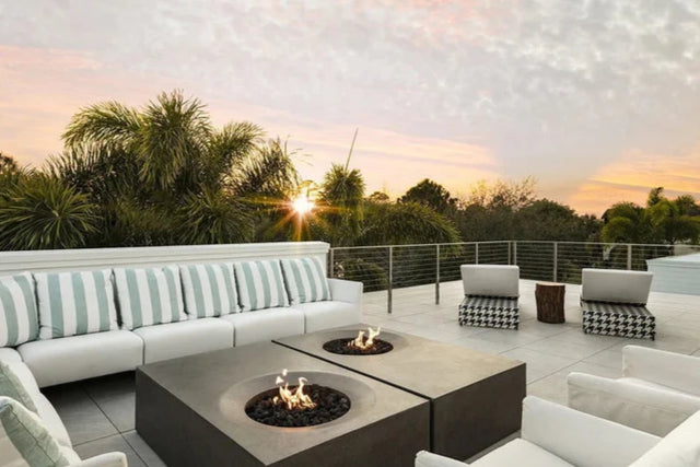 Luxury rooftop patio with two square gas fire pits, surrounded by white cushioned lounge seating and palm trees at sunset